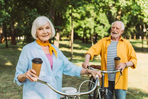 Selective focus of elderly woman holding paper cup near bike and husband in park — Stock Photo