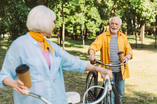 Enfoque selectivo del hombre mayor alegre sosteniendo la taza de papel cerca de la bicicleta y la esposa en el parque - foto de stock