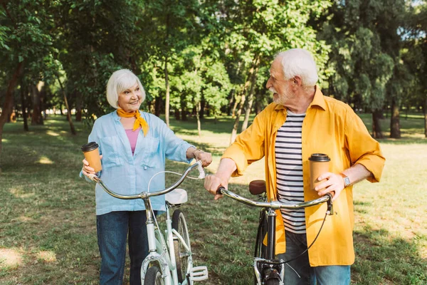 Fröhlicher Senior hält Coffee to go in der Hand und schaut Frau neben Fahrrad im Park an — Stockfoto