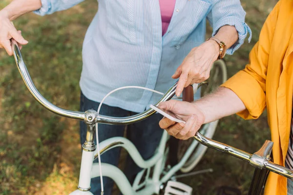 Vista ritagliata della donna anziana che punta con il dito allo smartphone vicino al marito con bici nel parco — Foto stock