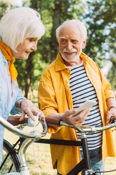 Enfoque selectivo del hombre mayor sosteniendo el teléfono inteligente cerca de la esposa sonriente y las bicicletas en el parque - foto de stock