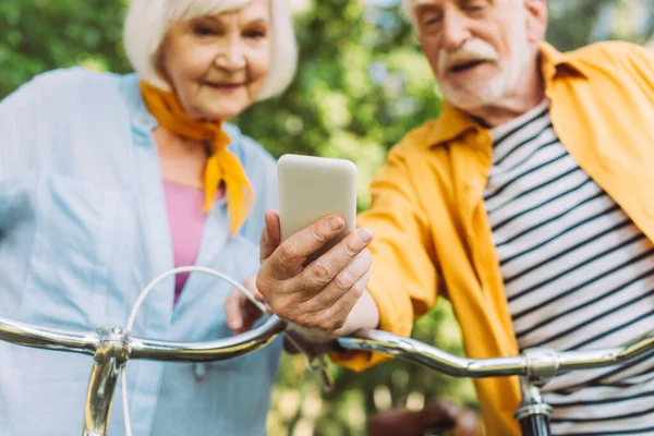 Foco seletivo do casal de idosos usando smartphone perto de bicicletas no parque — Fotografia de Stock