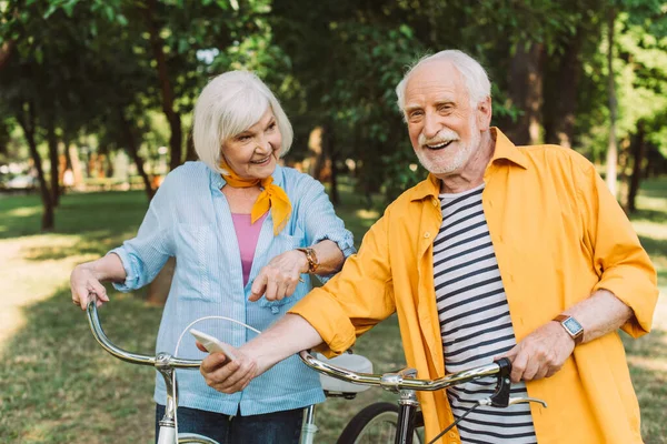Smiling elderly woman pointing with finger near husband with smartphone and bikes in park — Stock Photo