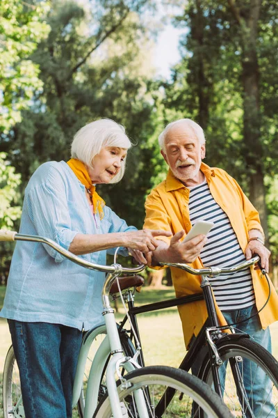 Seniorin zeigt auf Smartphone neben Ehemann und Fahrräder im Park — Stockfoto