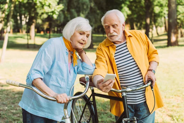 Elderly couple using smartphone near bikes in park — Stock Photo