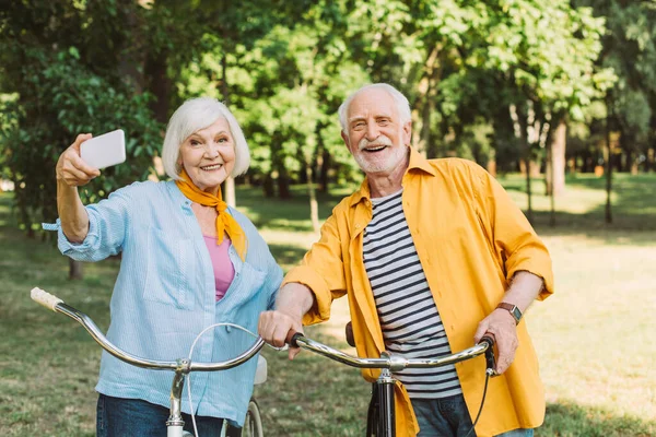 Alegre pareja de ancianos tomando selfie cerca de las bicicletas en el parque - foto de stock