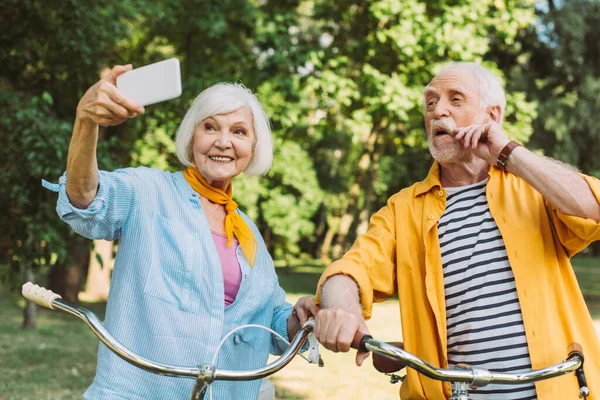 Старша жінка посміхається, беручи селфі біля чоловіка і велосипеди в парку — стокове фото