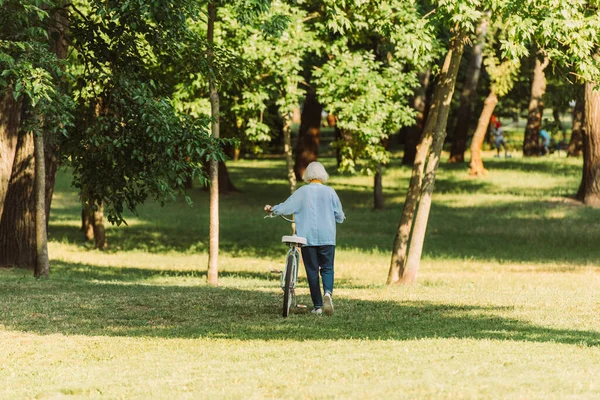 Visão traseira da mulher idosa andando com bicicleta na grama no parque — Fotografia de Stock