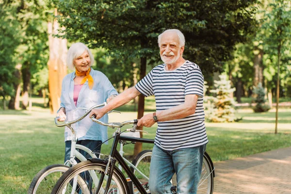 Улыбающийся мужчина смотрит в камеру во время прогулки с велосипедом рядом с женой в парке — стоковое фото