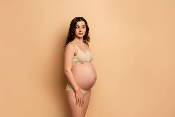 Mujer embarazada reflexiva en lencería mirando hacia otro lado mientras está de pie en beige - foto de stock