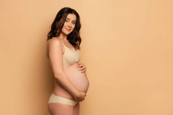 Brunette pregnant woman in underwear touching belly and looking at camera on beige — Stock Photo