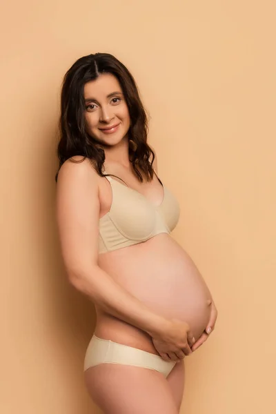 Pregnant brunette woman in lingerie looking at camera while touching belly on beige — Stock Photo