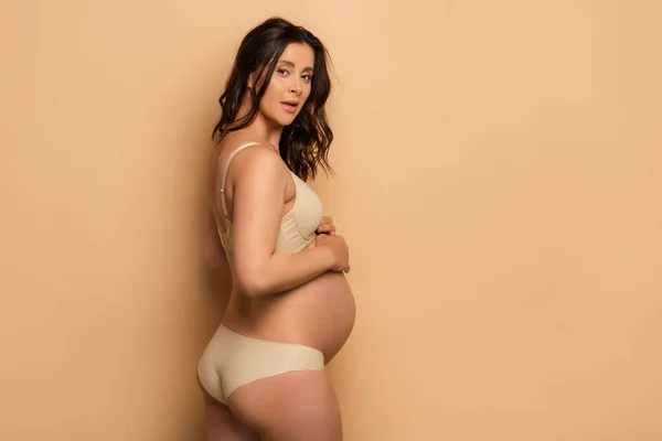 Pregnant brunette woman in underwear looking at camera while touching belly on beige — Stock Photo