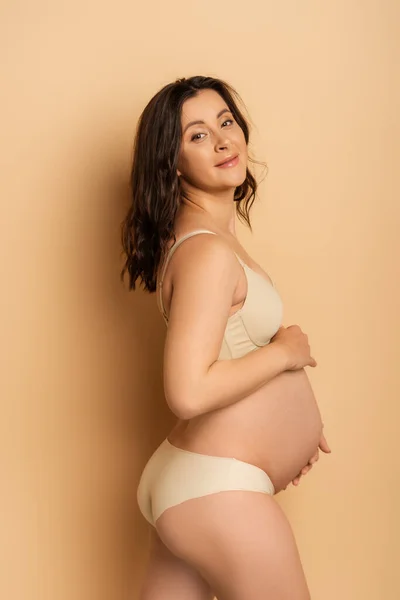 Брюнетка вагітна жінка в нижній білизні торкається живота і дивиться на камеру на бежевому — стокове фото