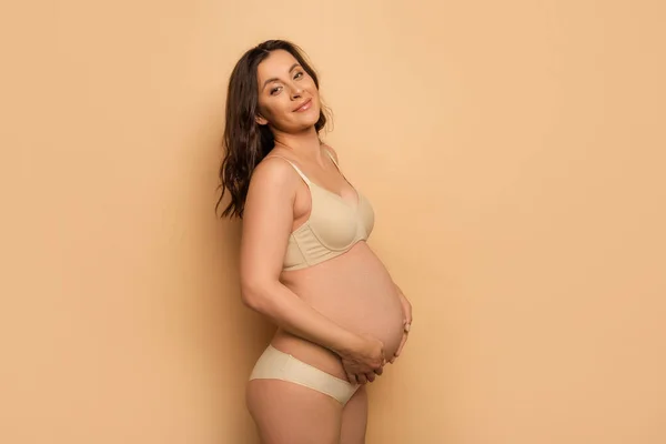 Pregnant brunette woman touching belly and looking at camera on beige — Stock Photo