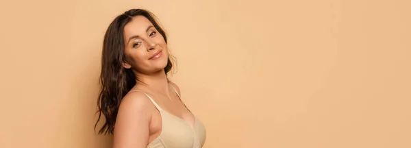 Panoramic shot of young brunette woman in bra looking at camera on beige — Stock Photo