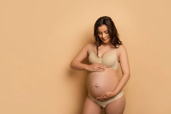 Jeune femme enceinte en sous-vêtements touchant le ventre sur beige — Photo de stock