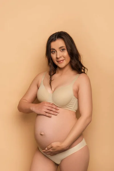 Pregnant brunette woman in lingerie touching belly and looking at camera on beige — Stock Photo