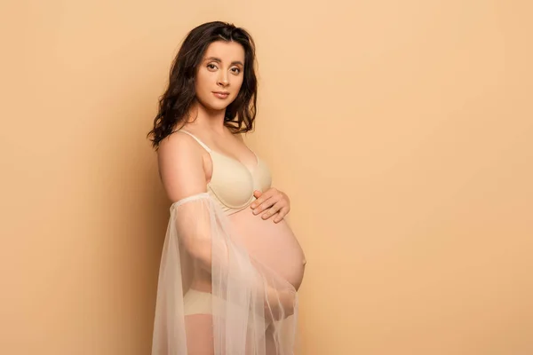 Pregnant woman in lingerie and chiffon sleeves touching belly and looking at camera on beige — Stock Photo