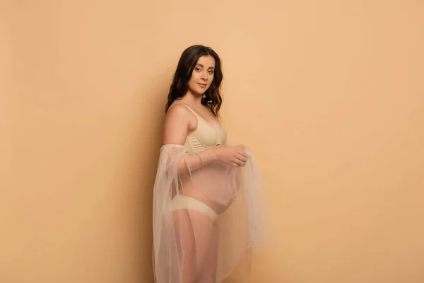 Morena mujer embarazada en lencería y mangas de gasa mirando a la cámara en beige - foto de stock