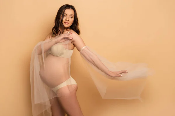 Young pregnant woman in underwear and chiffon sleeves posing on beige — Stock Photo