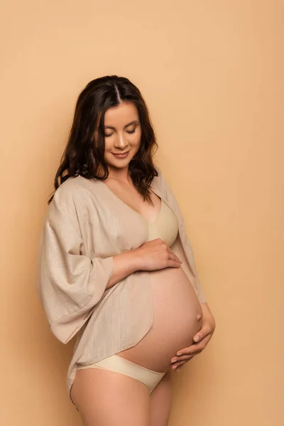 Young pregnant woman in lingerie and shirt touching tummy on beige — Stock Photo