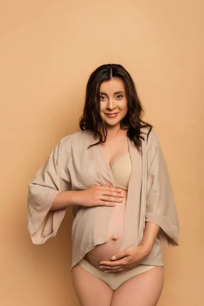 Young pregnant woman in shirt and lingerie touching belly and looking at camera on beige — Stock Photo