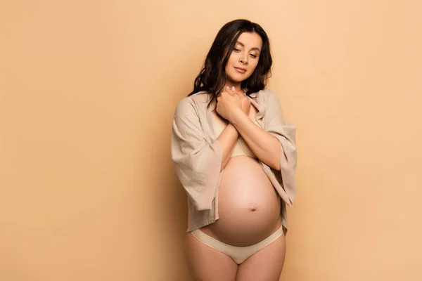 Mujer embarazada reflexiva en lencería y camisa con las manos apretadas cerca del pecho en beige - foto de stock