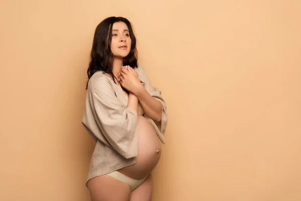 Tense pregnant woman in lingerie and shirt looking away while holding clenched hands near chest on beige — Stock Photo