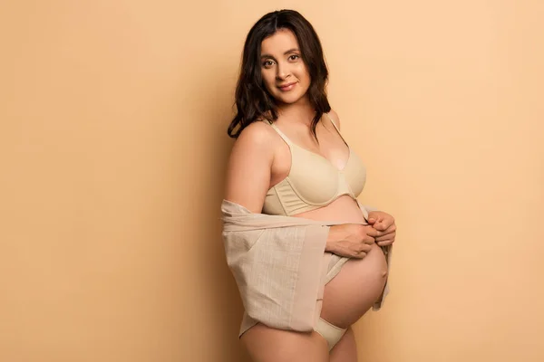 Brunette femme enceinte en lingerie couvrant ventre avec chemise sur beige — Photo de stock