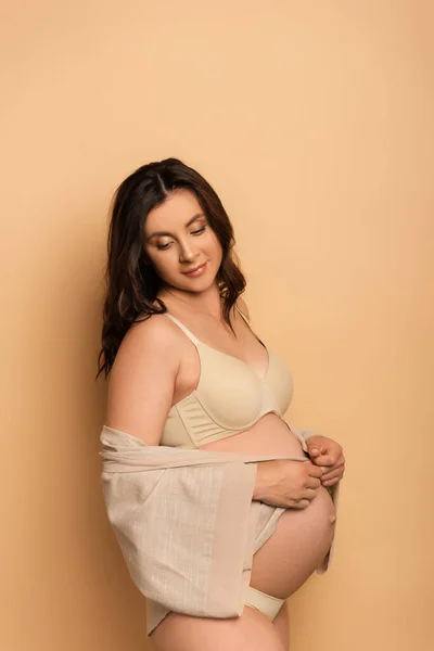 Young pregnant woman in underwear covering belly with shirt on beige — Stock Photo