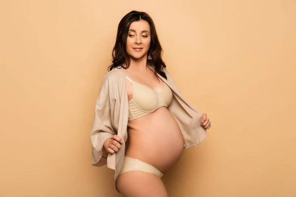Embarazada morena mujer en ropa interior tocando camisa mientras mira hacia otro lado en beige - foto de stock