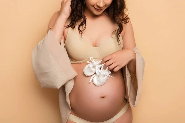 Ausgeschnittene Ansicht einer schwangeren Frau in Unterwäsche und Hemd mit Babystiefeln am Bauch auf Beige — Stockfoto