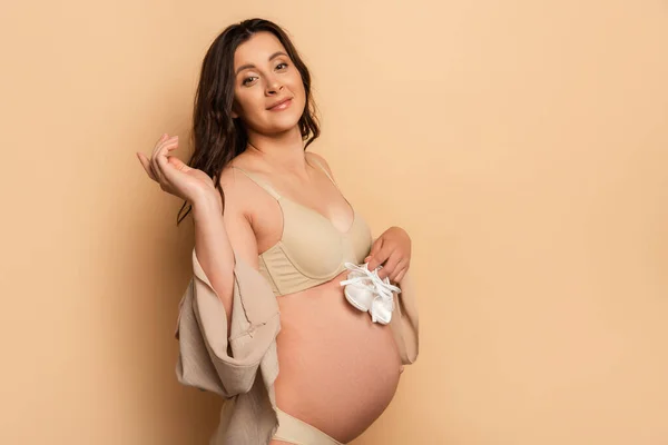 Молода вагітна жінка в нижній білизні тримає дитячі пінетки, дивлячись на камеру на бежевому — стокове фото