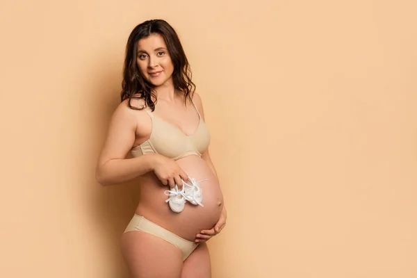 Jovem grávida tocando barriga, segurando botas de bebê e olhando para a câmera no bege — Fotografia de Stock