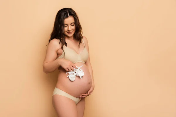 Brunette pregnant woman in underwear touching belly and holding baby booties on beige — Stock Photo