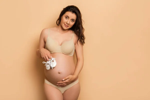 Jeune femme enceinte en lingerie tenant des chaussons de bébé et regardant la caméra sur beige — Photo de stock