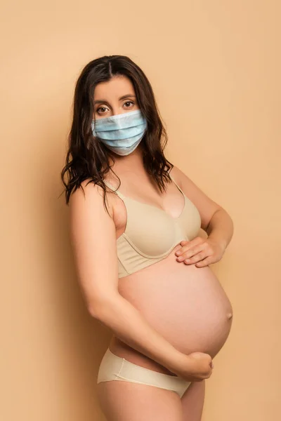 Mujer embarazada en lencería y máscara médica tocando la barriga y mirando a la cámara en beige - foto de stock