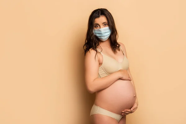 Jovem grávida em roupa interior e máscara médica tocando barriga e olhando para a câmera no bege — Fotografia de Stock