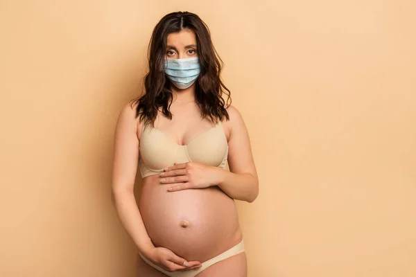 Morena, mulher grávida em máscara protetora tocando barriga e olhando para a câmera no bege — Fotografia de Stock
