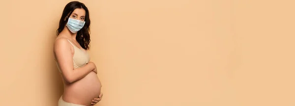 Panoramic crop of young, pregnant woman in medical mask touching tummy and looking at camera on beige — Stock Photo