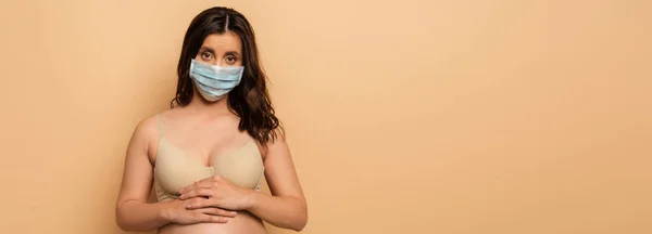 Horizontal image of brunette, pregnant  woman in protective mask touching tummy on beige — Stock Photo