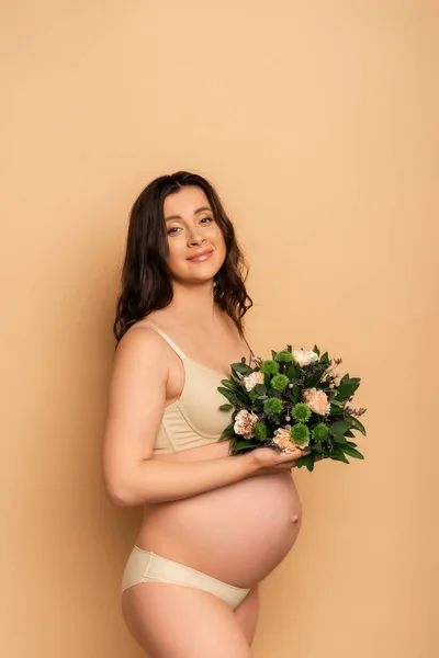 Brunette pregnant woman in lingerie holding bouquet of fresh flowers and looking at camera on beige — Stock Photo