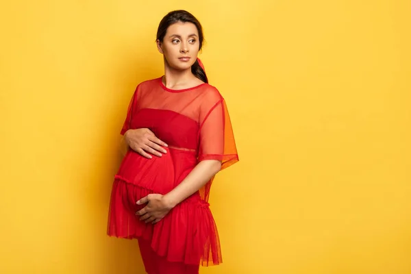 Thoughtful pregnant woman in red tunic touching tummy while looking away on yellow — Stock Photo