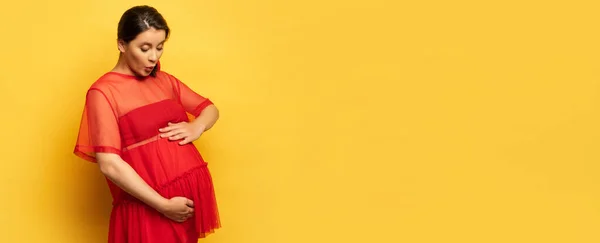 Заголовок веб-сайту шокованої, вагітної молодої жінки в червоній туніці, що торкається животик на жовтому — стокове фото
