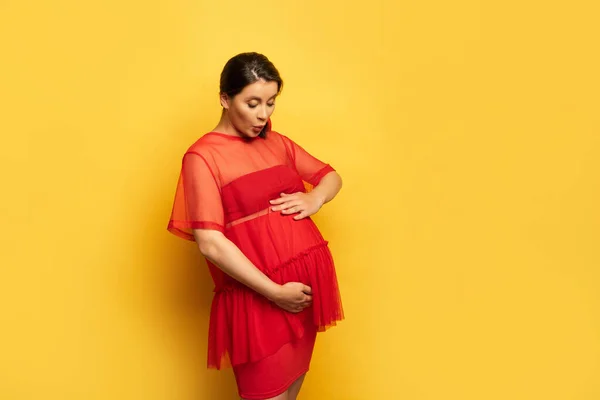 Chocado grávida jovem mulher em túnica vermelha tocando barriga no amarelo — Fotografia de Stock