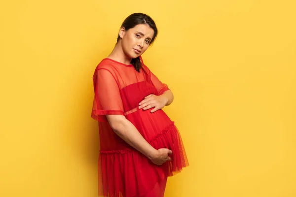 Pregnant young woman in red outfit worried while touching belly and looking at camera on yellow — Stock Photo