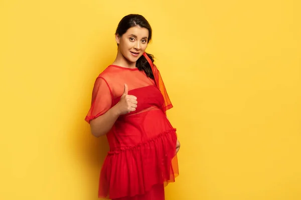 Mulher grávida em túnica vermelha mostrando polegar para cima enquanto olha para a câmera no amarelo — Fotografia de Stock