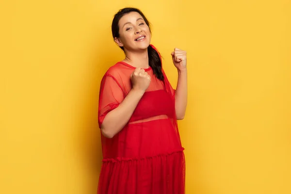 Emocionada mujer embarazada en traje rojo mostrando gesto ganador mientras mira a la cámara en amarillo - foto de stock