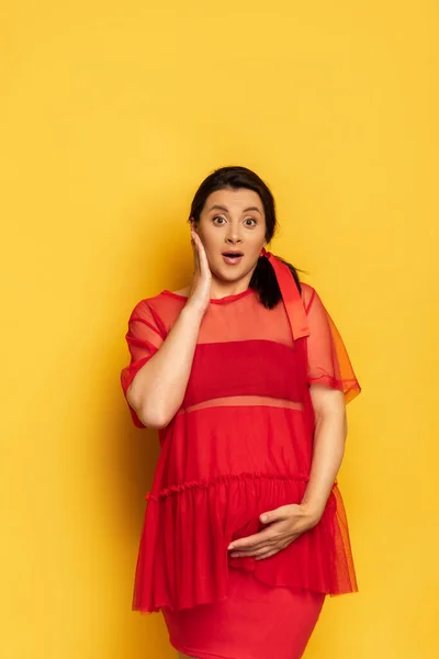 Excited pregnant woman in red tunic touching tummy and face while looking at camera on yellow — Stock Photo